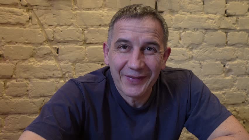 middle-aged man sitting against background brick wall, indoors, laughingly tells funny story to camera.