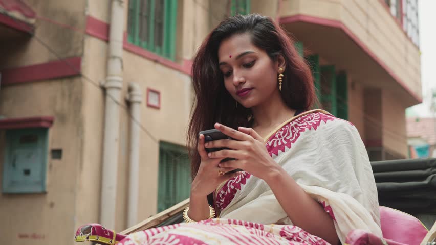 A young Indian female sitting in a moving local hand pulled auto rickshaw traveling through an old city street or lanes using a mobile phone to type a text message or SMS with a smile on her face.