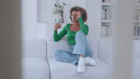 Young pretty African American woman listens to music in wireless headphones and dances sits on sofa holds phone in hands and watches clip or broadcast of concert located in spacious apartment.