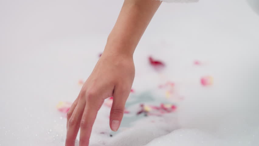 Lady dips hand into foam with rose petals closeup. Woman prepares romantic bath with fresh flowers stirring foam in bathtub. Idea for Valentine day