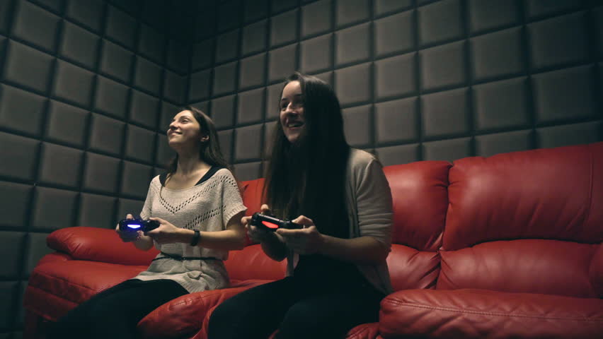 Two young girls playing video games with game controller on red sofa