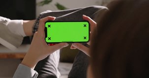 Happy young woman holding smartphone with green screen while resting on sofa couch. Watching movies on the phone. Advertisement of app. Watching video content. Modern technologies. Leisurely moments