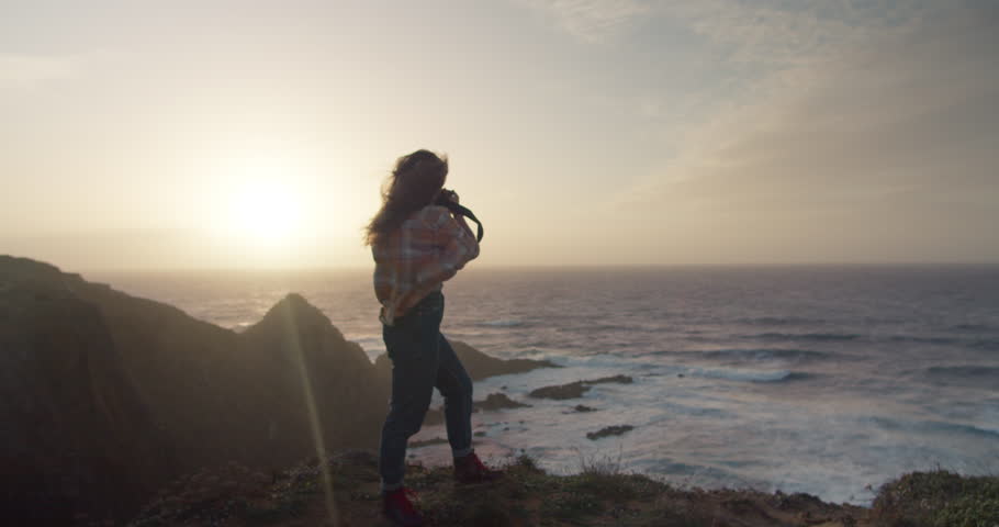 Epic and cinematic shot of female adventure or travel photographer stand on edge of cliff in evening sunset light. Wind blow her hair around. Explore exotic travel destination and create content Royalty-Free Stock Footage #3397464993