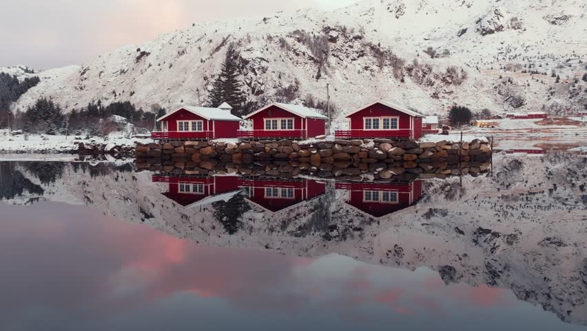 Iconic red rorbu houses in a traditional fishing village on Lofoten Islands, Norway, create a picturesque winter scene with snow-covered vistas. The serene beauty is mirrored in the calm water.