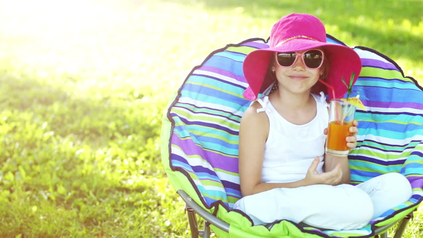 Happy child sitting in a chair outdoors in sunny day. Slow motion. Girl drinking
