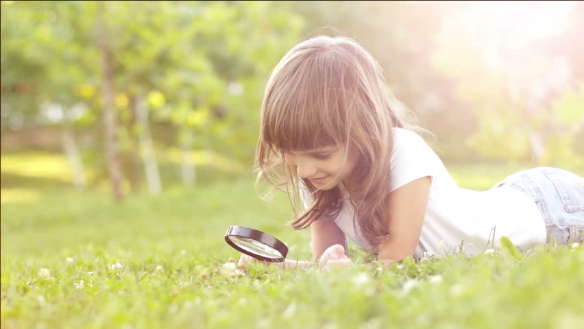 Portrait girl with magnifier lying on the grass outdoors in sun lights