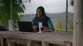 African American young boy sits in wooden gazebo and talks at video call using laptop and phone on tripod. Teenager remotely studies during holidays in mountain forest. Concept of outdoor studying.