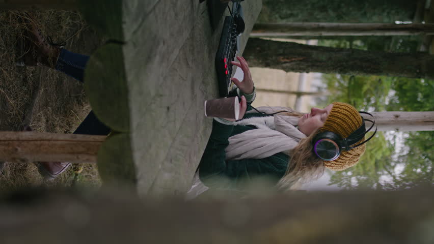 Caucasian woman wearing headphones plays on MIDI keyboard outdoors. Female musician creates and records music, mixes songs, uses laptop while sitting in gazebo in mountain forest. Vertical view.