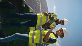 Vertical video of male and female engineers wearing hard hats and hi vis safety vests with digital tablet outdoors inspecting solar panels on sustainable energy farm - shot in slow motion