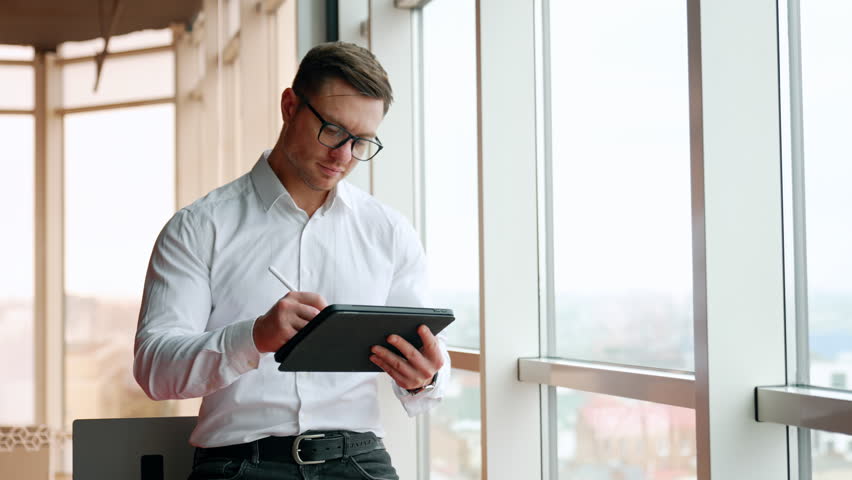 Busy male employee stands leaning on the desk and holding an I-pad. Businessman in eyeglasses drawing or making notes on I-pad.