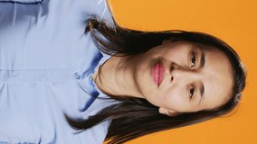 Vertical Video Filipino model praying for forgiveness and showing gratitude on camera, hopeful woman with belief in spirituality. Asian person with prayer hands having faith in God, orange background.