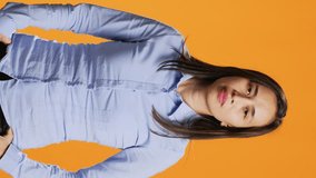 Vertical Video Portrait of carefree asian girl posing with confidence, wearing blue shirt and feeling confident in studio. Pretty filipino person standing over orange background, smiling with elegance