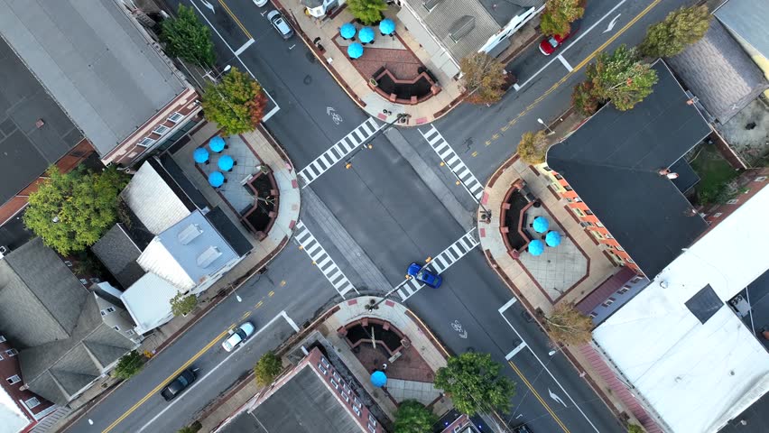 Crosswalks at intersection of town square. Blue umbrellas at an outdoor café on a city street. Aerial top down spinning shot as traffic drives. Royalty-Free Stock Footage #3398709785