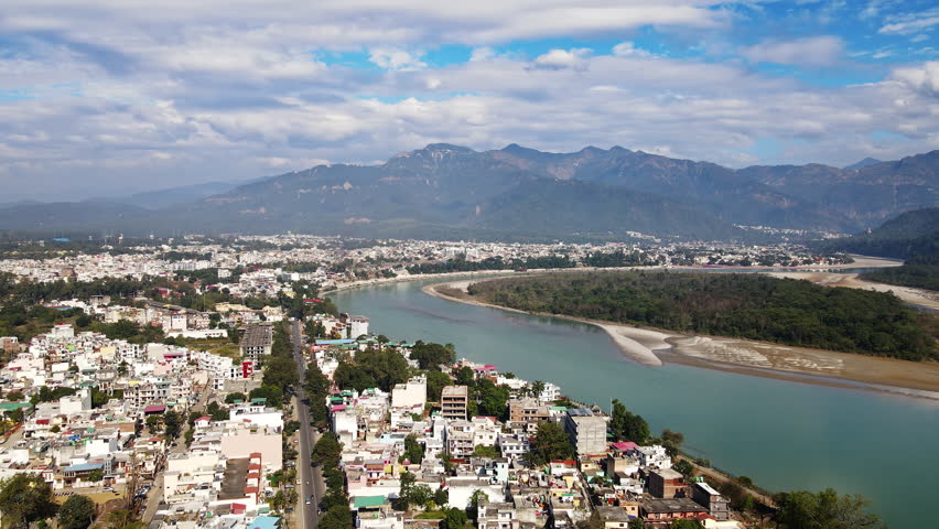 A bird's-eye view of Rishikesh City in Uttarakhand surrounded by lush green hills on a cloudy day. It is a hilly Indian town located along the Ganges River. Beautiful Indian city. Royalty-Free Stock Footage #3398775547