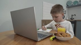Little boy is sitting at a table in the kitchen, eating liver, drinking juice, drawing in a notebook, watching videos on a laptop