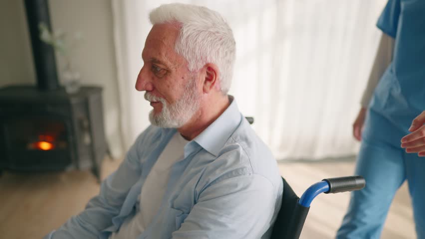 Old man in wheelchair talks with nurse. Senior paralyzed male thanks social worker woman for help support at home. Caregiver holds hand on patient shoulder stands near. Legs paralysis,   Royalty-Free Stock Footage #3398851745