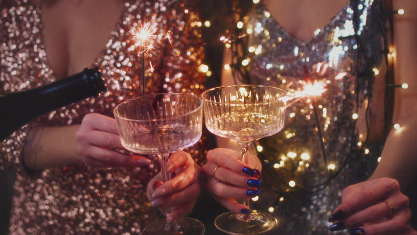 The girls pour champagne into round glasses from a bottle. Women in a sparkling dress. Festive decoration, Christmas garland. Full cocktail creamers - le coupe. The female