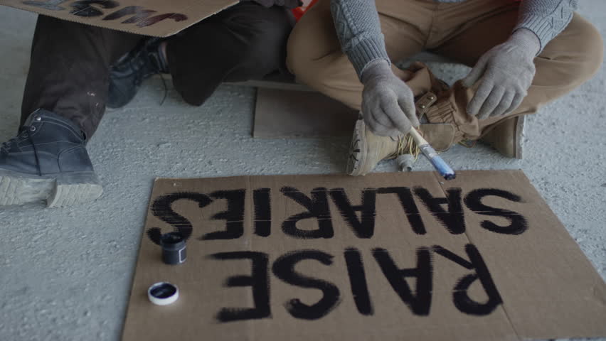 Tilting up shot of two ethnically diverse construction workers writing Raise Salaries and Justice Now slogans on pieces of cardboard, making protest banners before going on demonstration Royalty-Free Stock Footage #3398937327