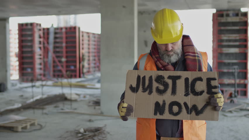 Medium slowmo portrait of serious middle-aged Caucasian male construction workers standing at construction site with cardboard Justice Now strike banner Royalty-Free Stock Footage #3398947217