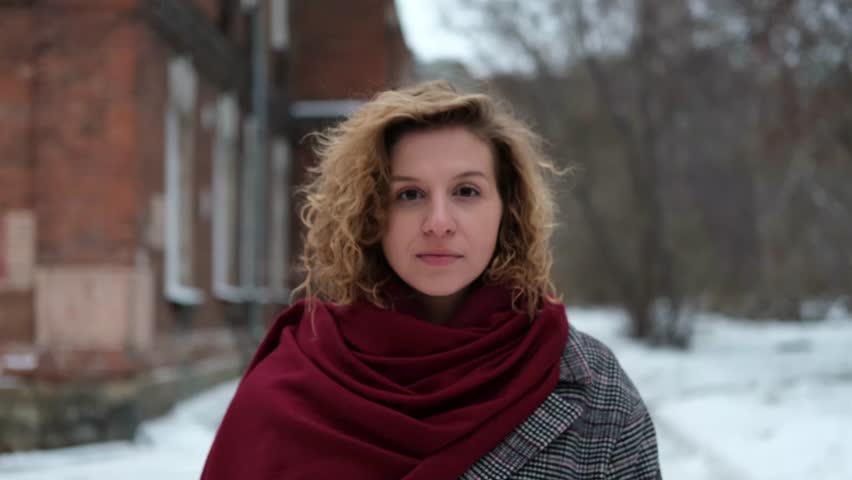 A young woman with curly hair is wearing a winter coat and a burgundy scarf. Woman on the street on a winter day. There