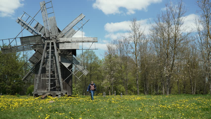 Focused preteen child running away from abandoned mill, handheld shot. Ethnic landscape with old wooden mill in yellow blooming dandelion field on sunny spring day. Royalty-Free Stock Footage #3399040183