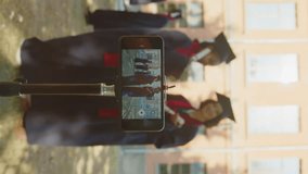 Vertical shot with selective focus on smartphone in tripod, group of students filming video during graduation ceremony outdoors
