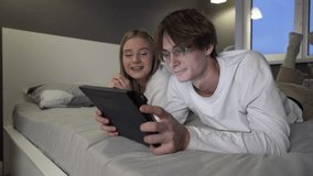 Couple is lying on the bed in the bedroom and uses a tablet to watch a video, a movie. Man, woman use touch screen for typing, social media apps or collaboration at home