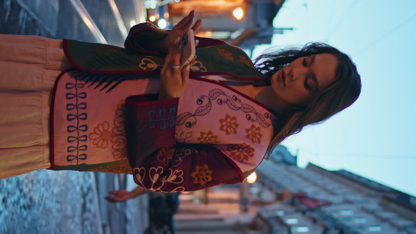 Girl calling taxi at night street using smartphone app. Serious woman tourist looking to cab at evening road traffic opening cellphone application vertical video. Stylish lady standing urban pavement.