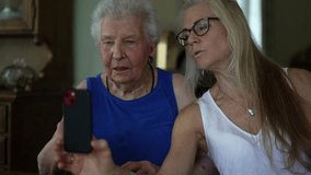Elderly senior woman and mature daughter having fun together looking at smartphone phone in dining room in slow motion.