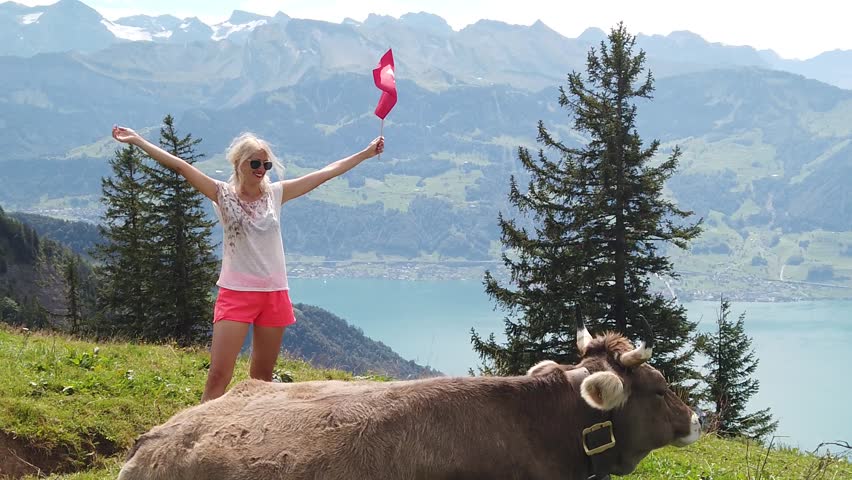 SLOW MOTION: girl with Swiss flag by a cow in alpine meadow along Rigi-Scheidegg railway with spectacular views of Swiss Alps, Schwyz basin, Lake Lucerne. Tourism in Lucerne area, Central Switzerland. Royalty-Free Stock Footage #3399360729