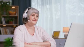 smiling female pensioner with gray hair talking on a video call on a laptop using a headset while sitting at table in a cozy room