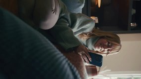 Vertical video of woman wearing cosy jumper sitting on sofa at home looking at mobile phone and drinking hot drink from cup - shot in slow motion
