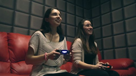 Two young girls playing video games with game controller on red sofa