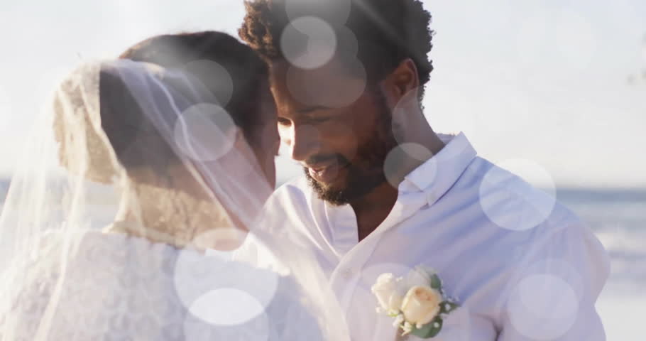 Animation of light spots over happy african american groom unveiling bride on beach at wedding. Romance, love, vacations and marriage, digitally generated video.