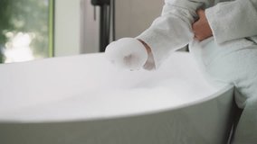 Woman takes foam sitting on bathtub edge. Young lady in soft bathrobe plays with bubble bath preparing for body care procedures at home. Home relaxation - Powered by Shutterstock - Get 15% off with code: PIKWIZARD15