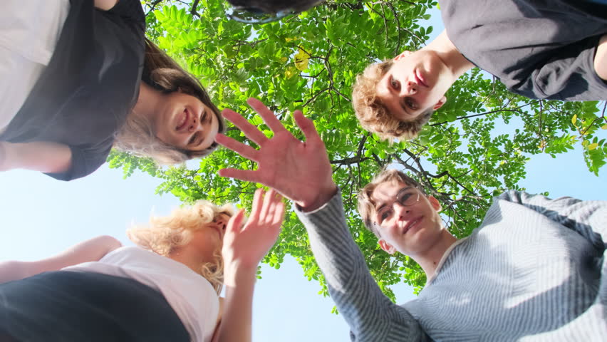 Happy students stand in circle under tree in park. Friends laugh in sunlight. Teenagers press down hands and raise up to sky Royalty-Free Stock Footage #3399724955