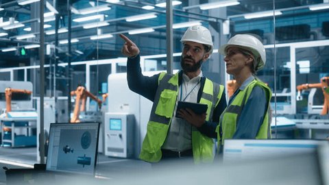 Hispanic Male Process Engineer Using Tablet And Talking To Caucasian Female Supervisor At Autonomous Assembly Line With Robotic Arms. Colleagues Wearing Hardhats, Discussing New Engine. : vidéo de stock