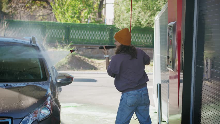 A young Caucasian woman washes her car at a self-service car wash on a sunny autumn day.