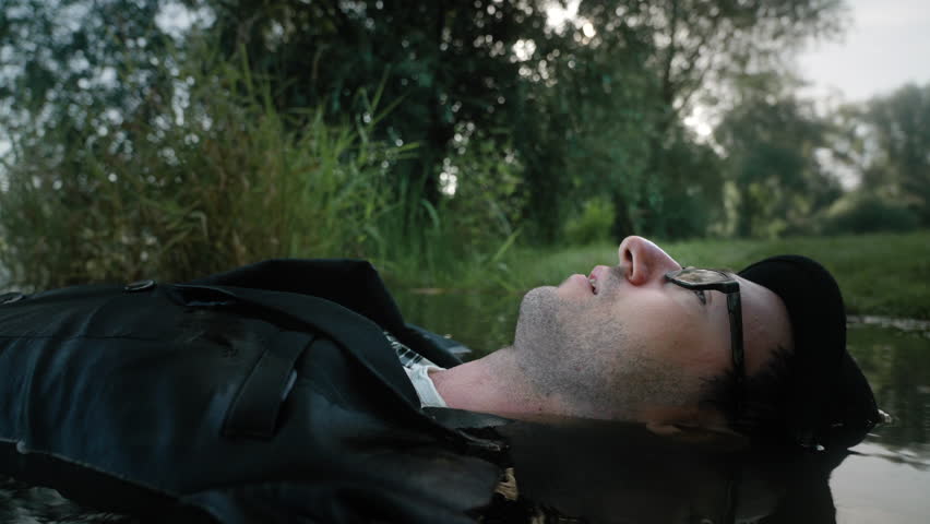 A closeup portrait footage of adult Caucasian man lies with his full clothes in the shallow lake water in the park in daylight