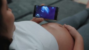 Last trimester pregnancy. Pregnant Woman Looking Ultrasound Video  Baby on mobile phone. Girl looking at Ultrasound vdo scan on smartphone lying on couch at home. Gynecology, Birth, Childbirth.