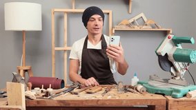 Young carpenter wearing brown apron and cap in his joinery holding smartphone broadcasting livestream in his joinery vlog talking about woodwork creating content about his profession