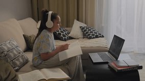 Full shot of young girl sitting on couch in her living room talking with her teacher while having online class