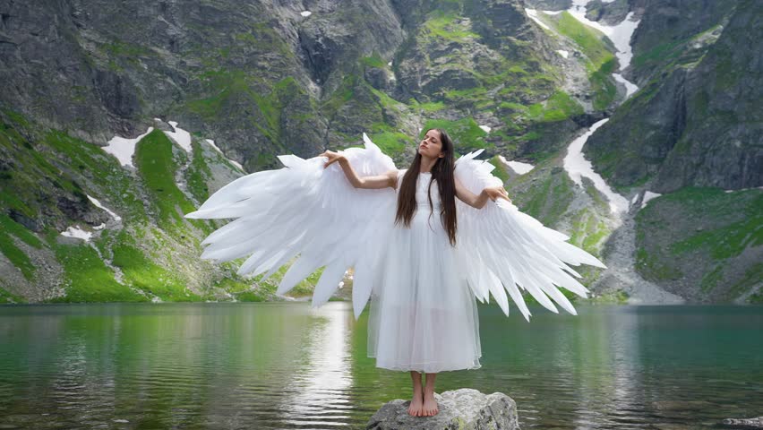 White angel with big wings on a beautiful mountain lake. A young long-haired girl in the image of an angel flaps her wings against a mountain 