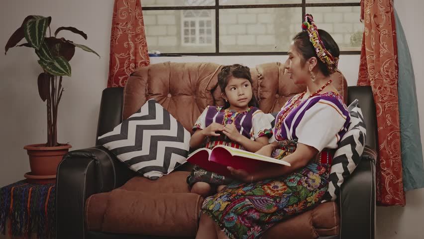 Mother reads a story to her daughter, both sitting on a sofa at home. Royalty-Free Stock Footage #3400183201