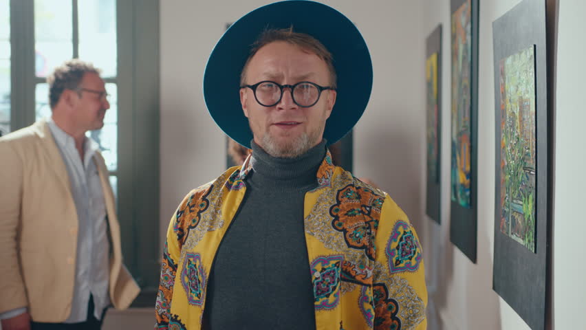 Stylish male artist wearing blue hat and bright shirt standing in exhibition hall of art gallery and posing for camera with a smile, people walking in the background. Medium shot, video portrait