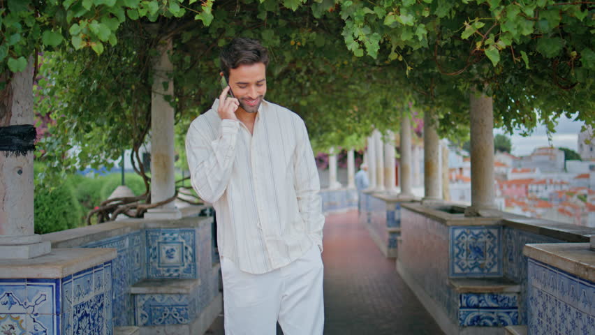 Relaxed brunette ending call mobile phone strolling alley. Happy gentleman hiding smartphone at pocket walking at cityscape boulevard. Smiling handsome man waiting girlfriend under green trees alone