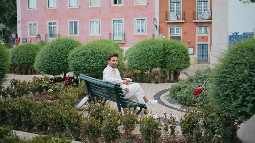 Romantic man waiting girlfriend at green park bench. Relaxed spanish boyfriend holding flower enjoying beautiful city at date place. Handsome bearded model relaxing at summer botanical garden alone 