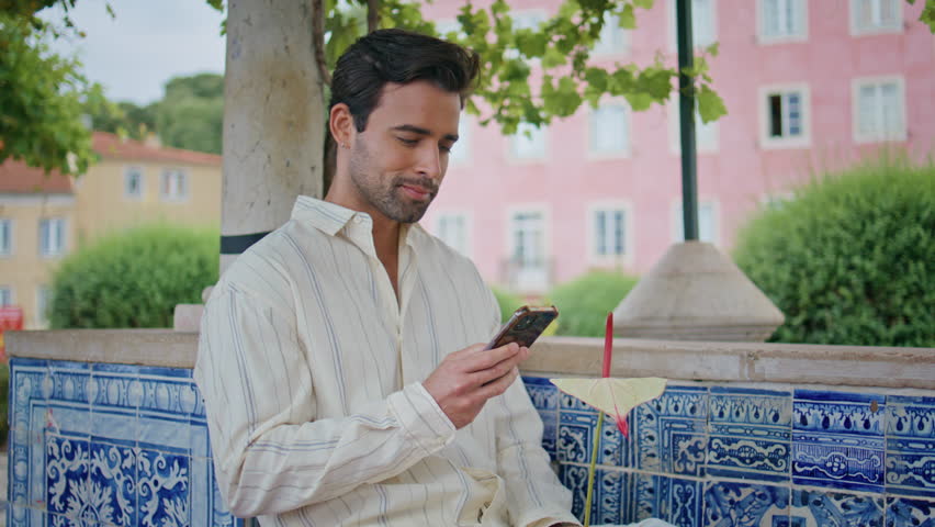 Happy macho calling smartphone waiting date at city park zoom on. Smiling hispanic man talking mobile phone standing alone at blue tile place closeup. Handsome brunette using cellphone outside Royalty-Free Stock Footage #3400220267