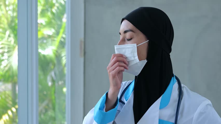 Muslim woman doctor at window takes off medical mask and rubs temple. Muslim woman doctor in black hijab sighs, pulls off medical mask and wearily rubs temple. Concept: tired doctor needs rest Royalty-Free Stock Footage #3400403495