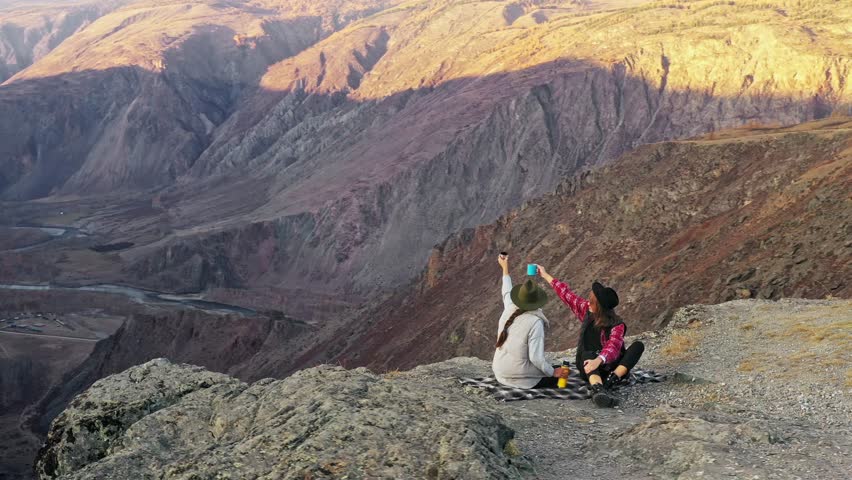 Two traveler female friends sitting on the edge of a sheer cliff above a deep cliff and chatting cheerfully drinking freshly brewed coffee watching the beauty of nature mountain forest and sunset on